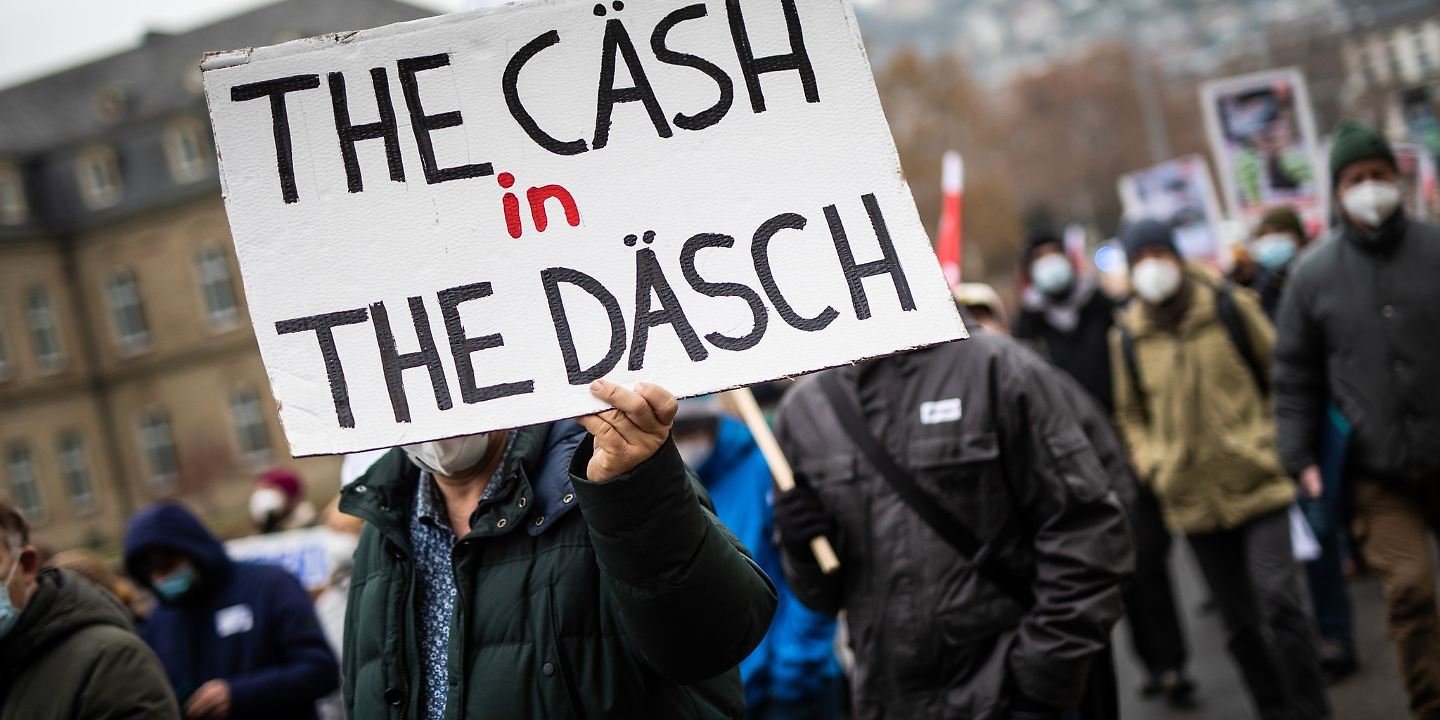 "The Cäsh in The Däsch" steht bei einer Kundgebung auf dem Schild eines Teilnehmers geschrieben.